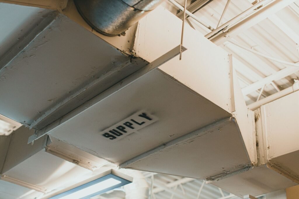 Rectangular white sheet-metal supply ducts running across an industrial ceiling with the word SUPPLY stencilled on one face, the kind of long duct run a dryer vent booster fan is fitted into