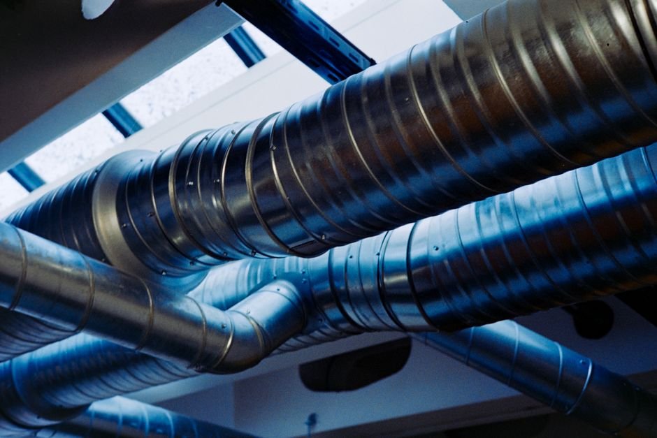 Spiral-wound rigid metal ducting with crimped joints and swept elbows running beneath a ceiling, lit blue from a window behind