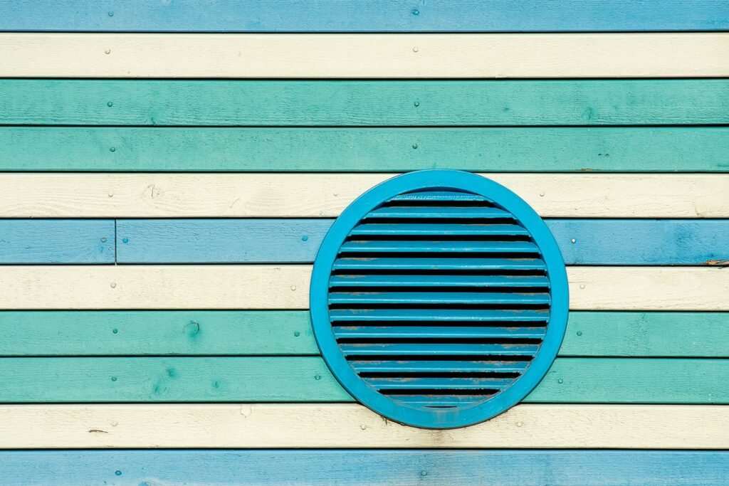 A round blue louvred wall vent fitted into painted horizontal wood siding, the fixed-louvre style of exterior termination a dryer vent wall cap should not be confused with