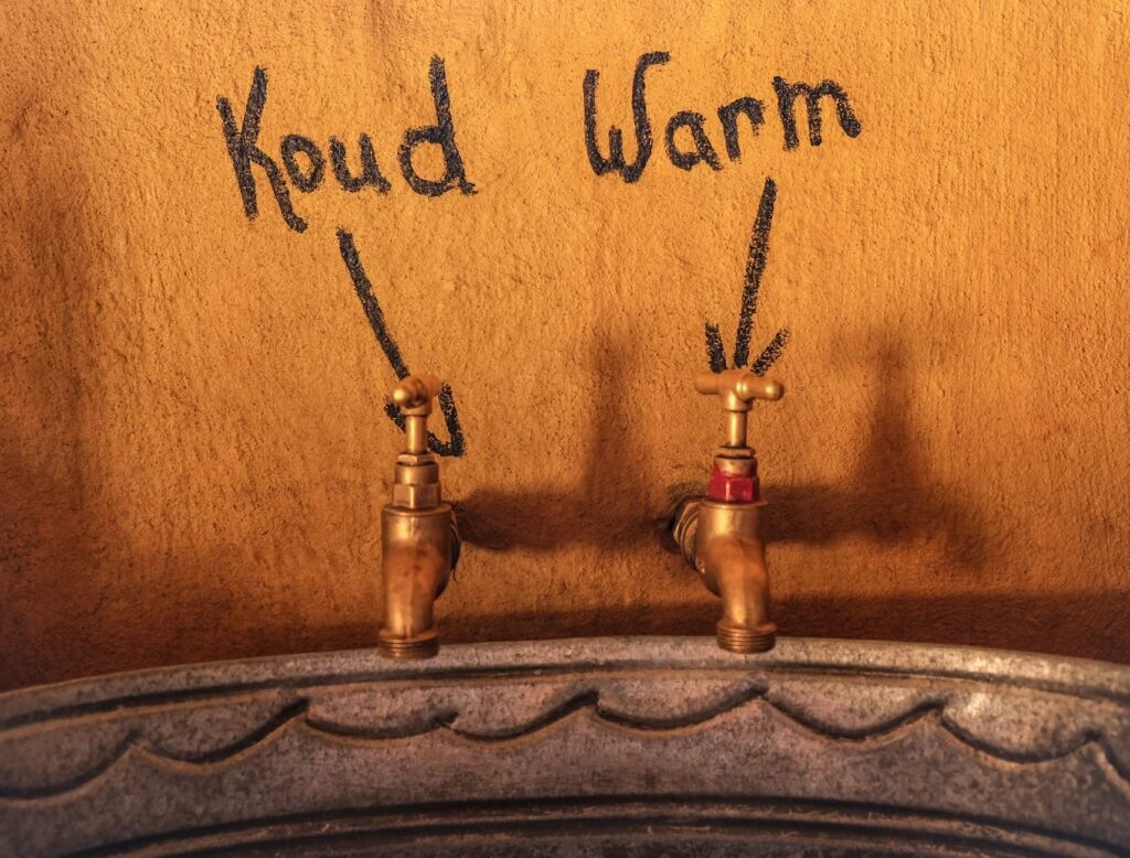 Two brass taps on an ochre plaster wall above a galvanised trough, with the words Koud and Warm chalked above them and an arrow drawn down to each tap