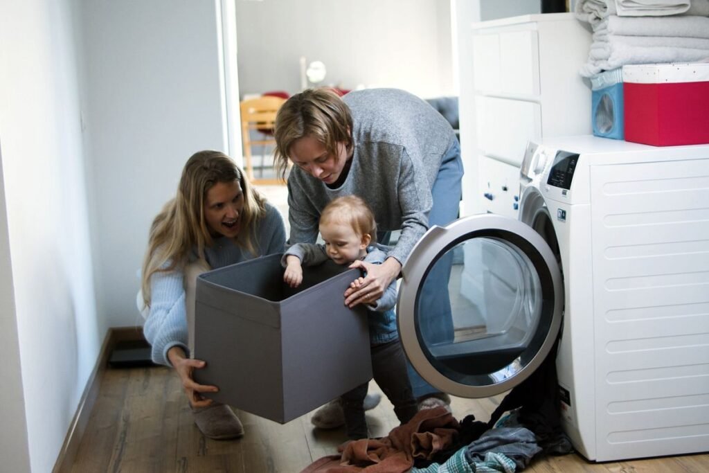 Two adults and a toddler crouched at an open front-load washer with a grey laundry box and clothes spread on the wooden floor