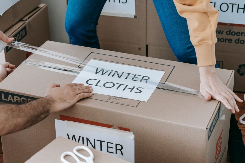 Two people taping shut a large cardboard carton hand-labelled for winter clothes, with more labelled boxes stacked behind them