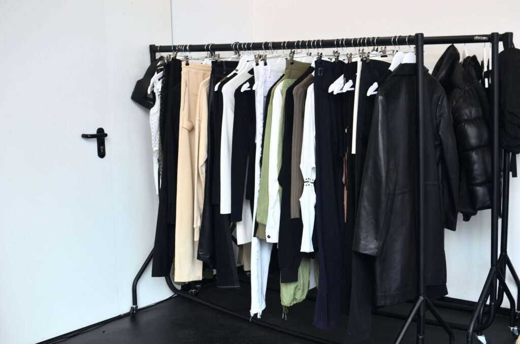 A black steel freestanding clothes rail packed tightly with black, cream and olive garments against a white wall beside a closed white door