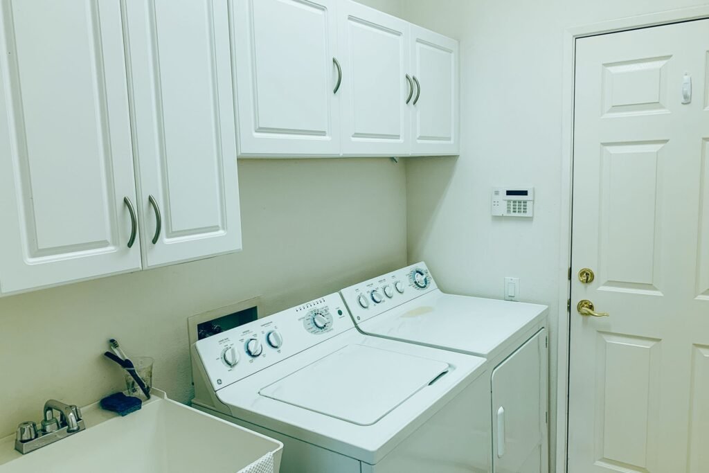 A white top-load washer and matching dryer with dial controls side by side under white cabinets in a tiled laundry room