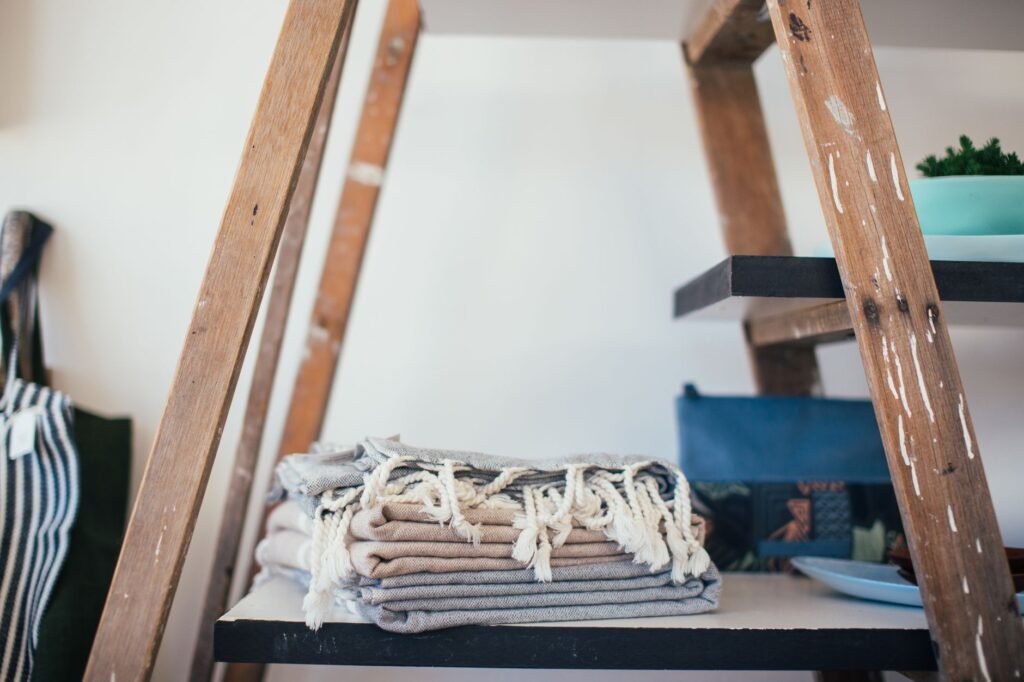 A stack of folded grey and beige cotton towels with knotted tassels on a painted wooden shelf, a stepladder frame and striped tote bag around them