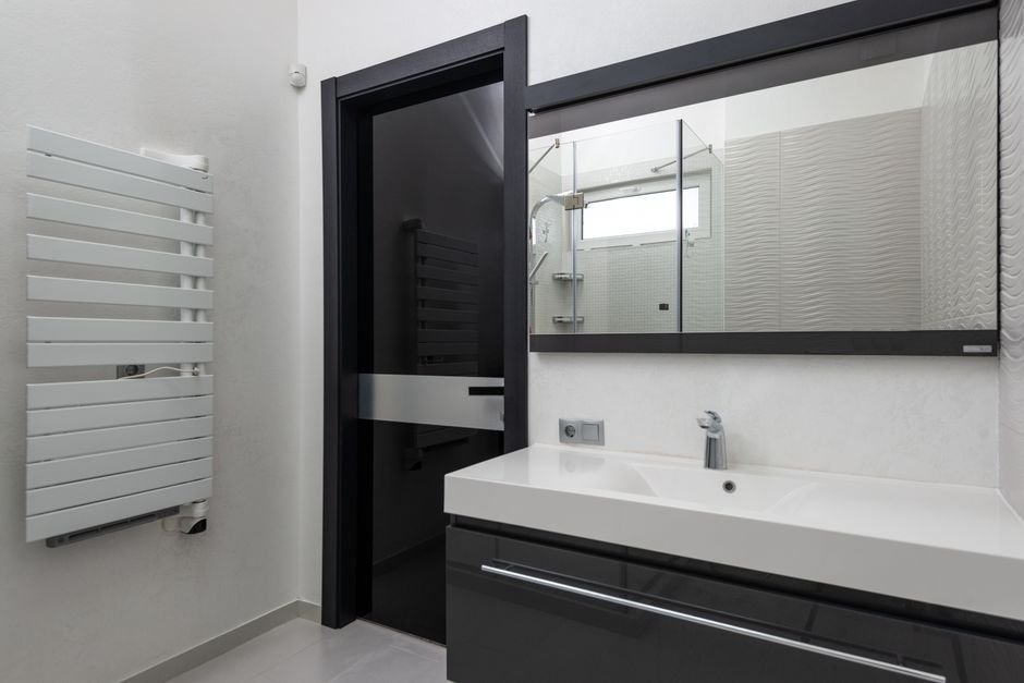 White flat-panel heated towel rail mounted on the wall of a grey and white bathroom beside an open dark door, with a second black rail visible in the room beyond