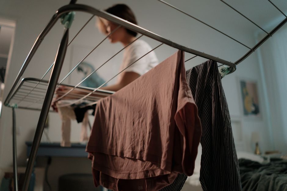 Close-up of a chrome folding clothes airer in a bedroom with a dusty pink T-shirt and striped trousers hung over the bars and a person loading it behind