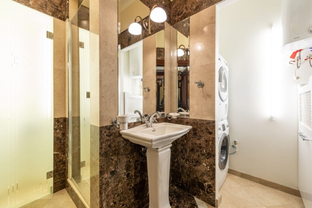 A white dryer stacked on a front-load washer in an alcove of a marble-tiled bathroom beside a pedestal sink and shower, an install with no exterior wall to duct into