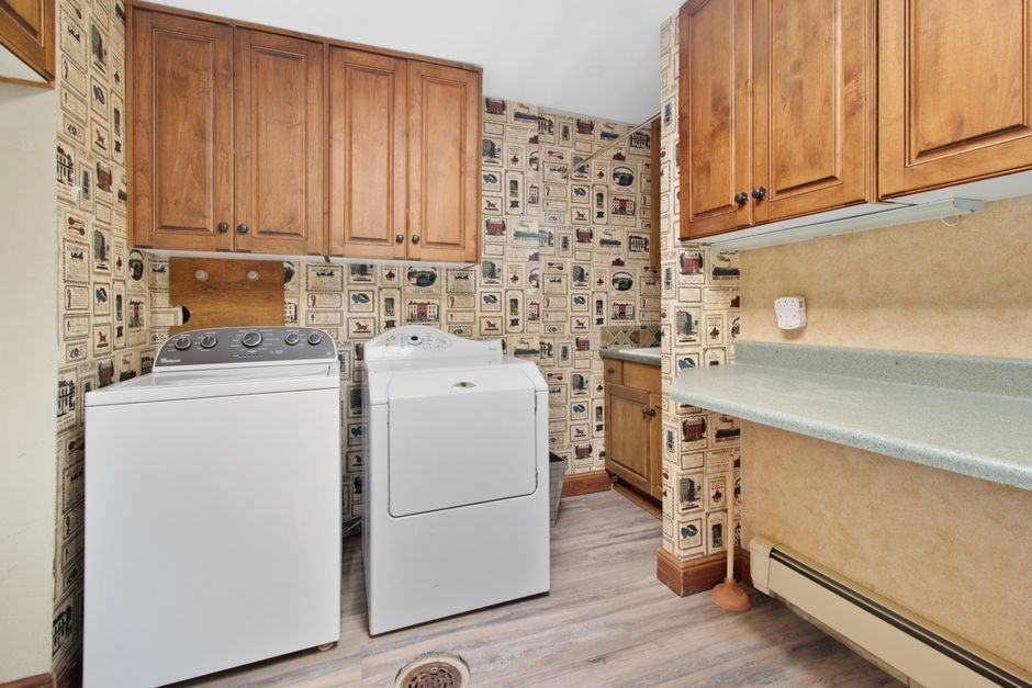 Home laundry room with a white top-load washer beside a white front-load dryer, wooden wall cabinets above and a floor drain in the vinyl