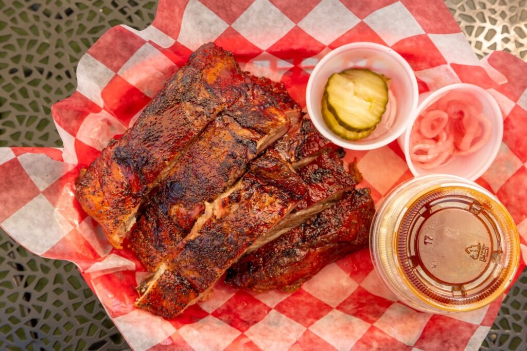 Overhead view of sliced, heavily glazed and charred barbecue ribs in red and white checked paper, with a tub of dark sauce, pickle slices and pickled onions beside them