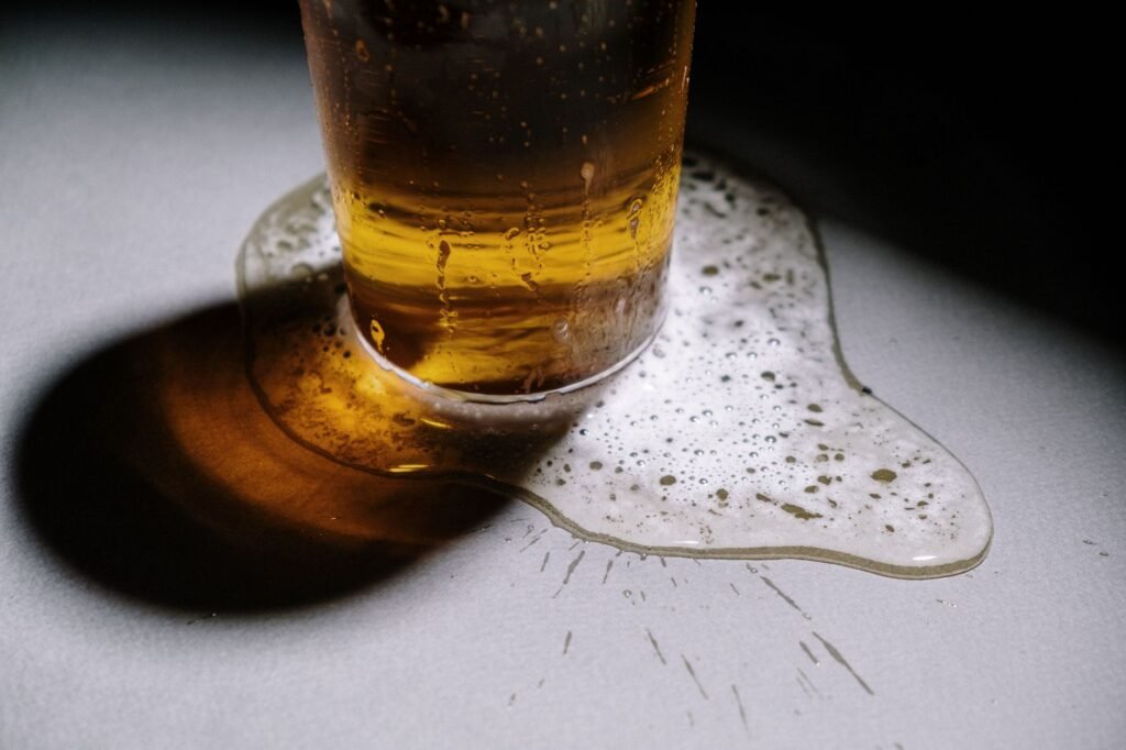 An overfilled pint glass of pale beer sitting in a wide puddle of beer and foam spreading across a grey surface