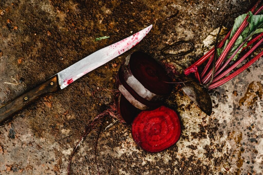 A kitchen knife with a deep red-stained blade lying on dark rusted stone beside a halved raw beetroot and a sliced round showing concentric magenta rings, with crimson stems and leaves alongside