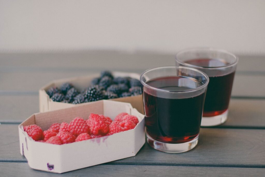 Cardboard punnets of raspberries and blackberries on a grey wooden table, the raspberry punnet stained purple where the juice has soaked in, beside two glasses of dark berry juice