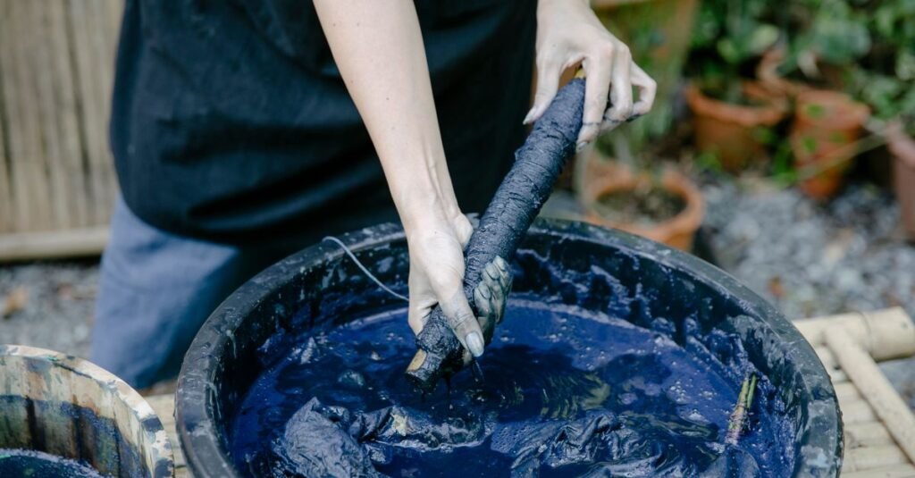 Dye-stained hands lifting a bound bundle of cloth out of a tub of deep indigo dye outdoors, the only real way to put colour back into fabric