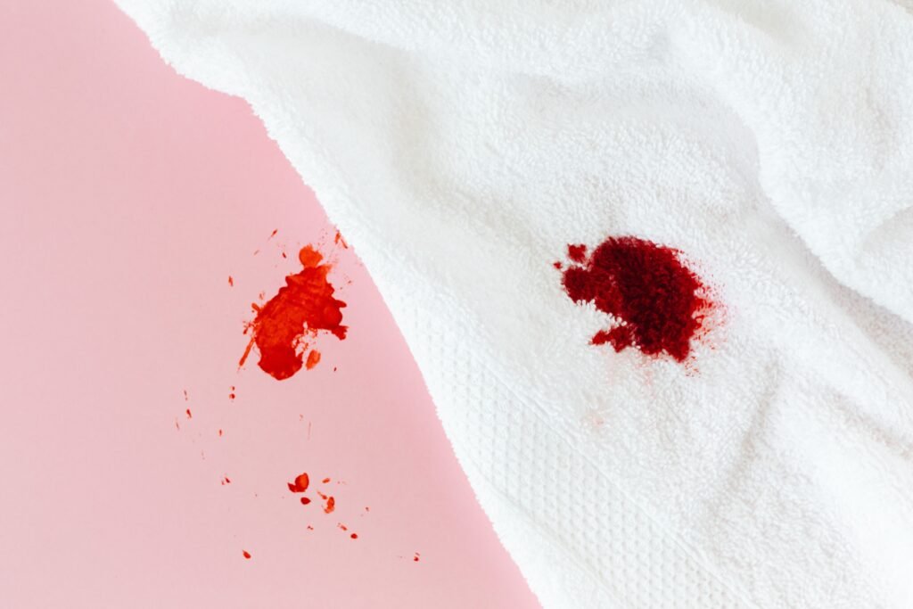 A fresh red blood spot soaked into a folded white cotton towel, with scattered droplets on the pink surface beside it