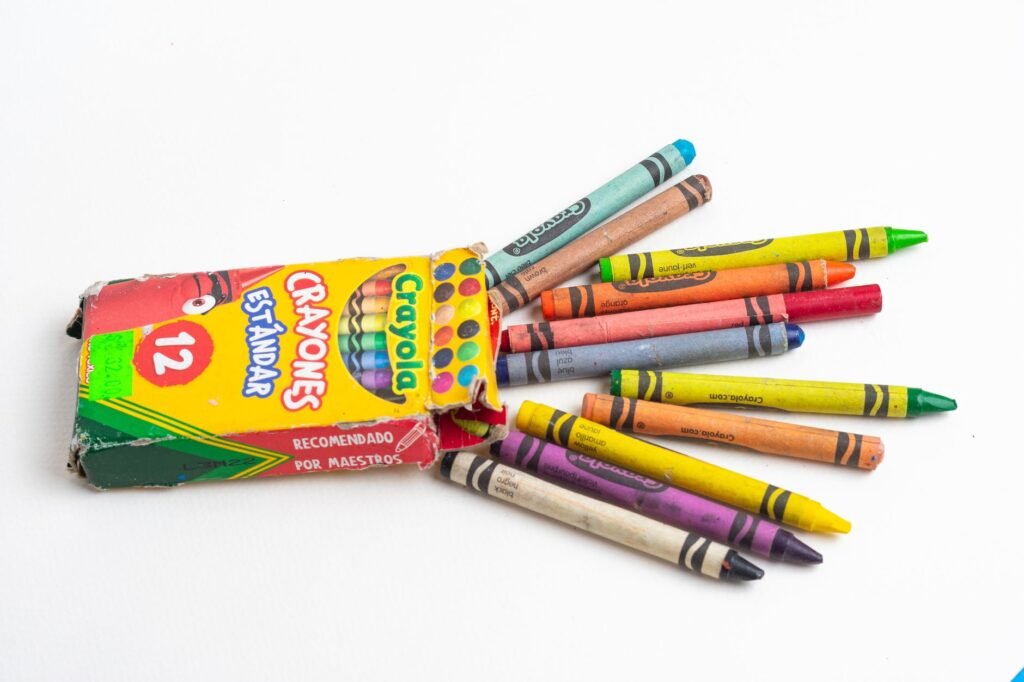 A battered Crayola crayon box lying open on a white surface with twelve coloured wax crayons spilling out of the torn end