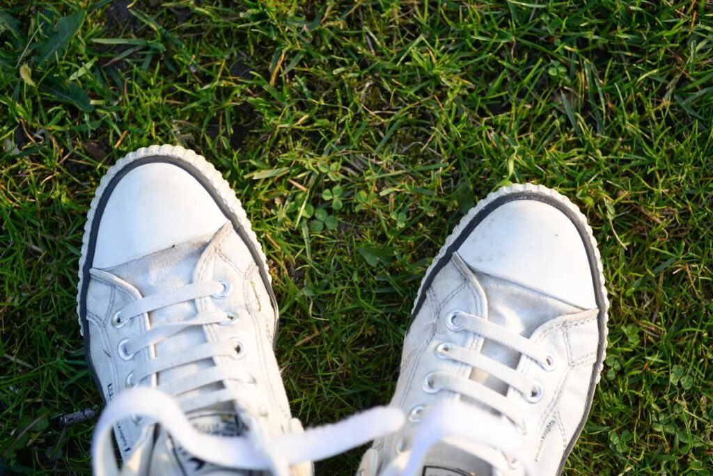 A pair of scuffed white canvas sneakers with white rubber toe caps standing on grass and clover, seen from above
