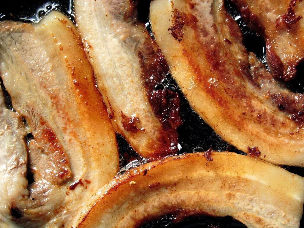 Close-up of fatty pork belly strips frying in their own rendered fat in a hot pan, the usual source of a grease stain on clothing