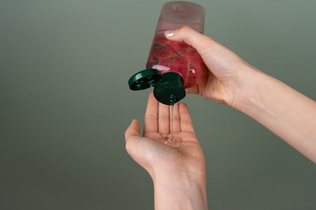 A clear gel being squeezed from a pink bottle with a green flip-top cap into an open palm against a plain olive-green background