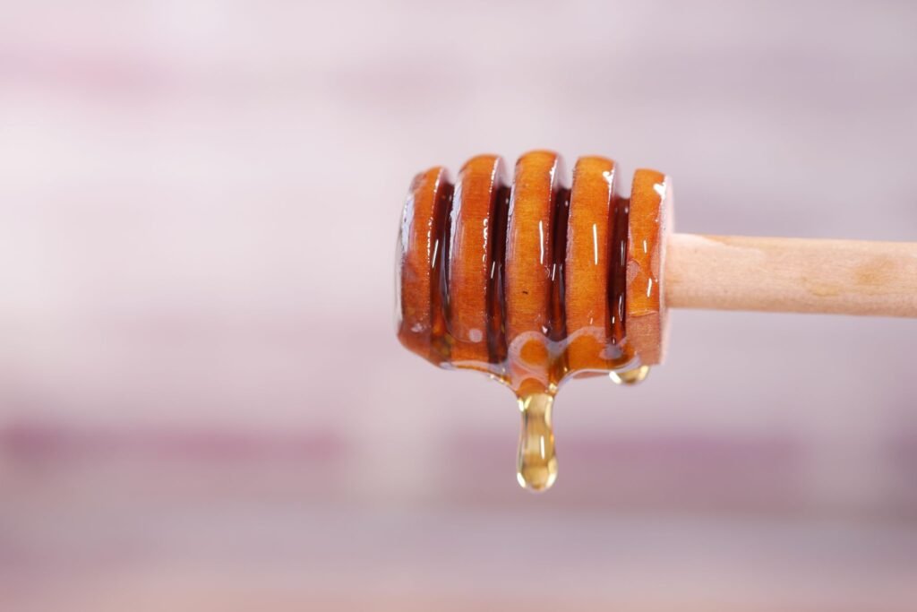 A wooden honey dipper held sideways with a single golden bead of honey hanging from its tip, about to fall, against a soft pink blurred background