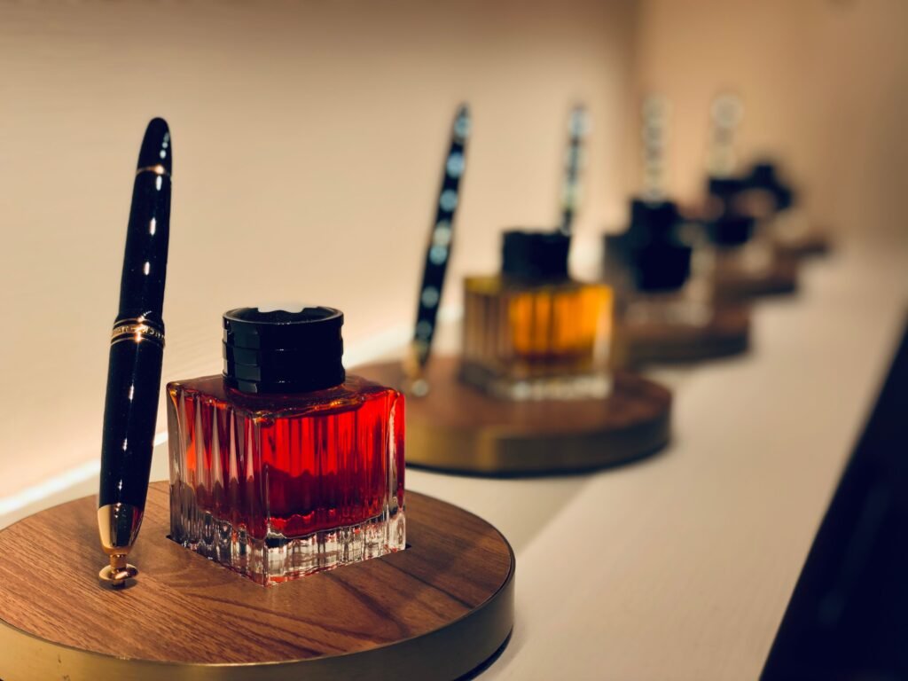 A black fountain pen standing beside an open bottle of deep red ink on a wooden stand, with more pens and ink bottles receding behind