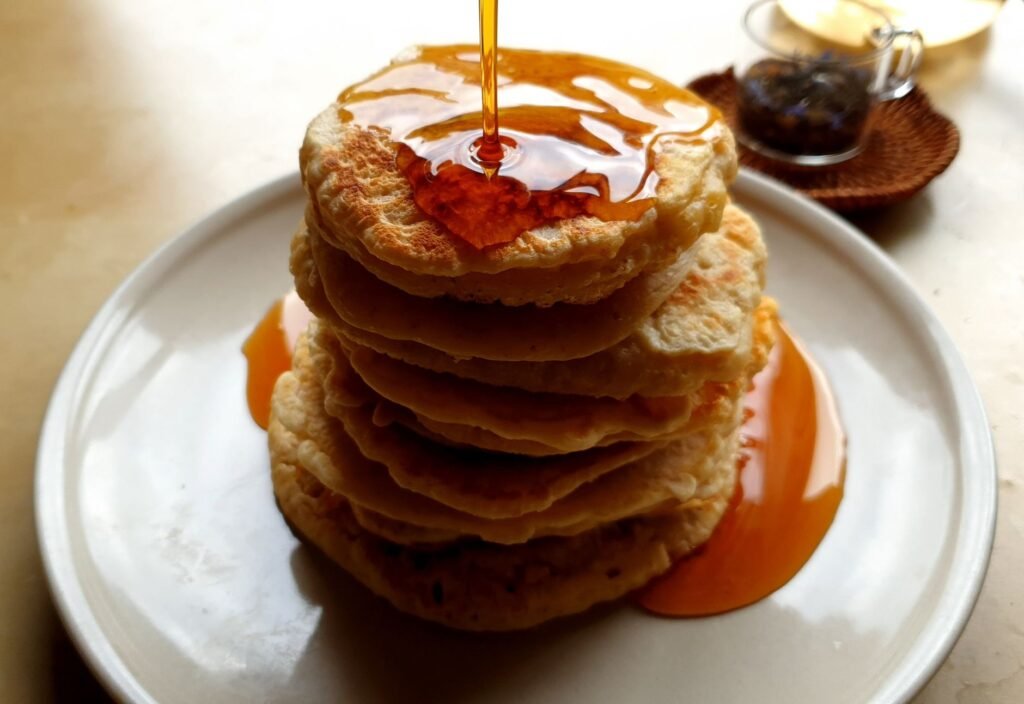 A thin stream of amber syrup being poured onto a tall stack of pancakes on a white plate, pooling on top and running down over the edge of the stack onto the plate