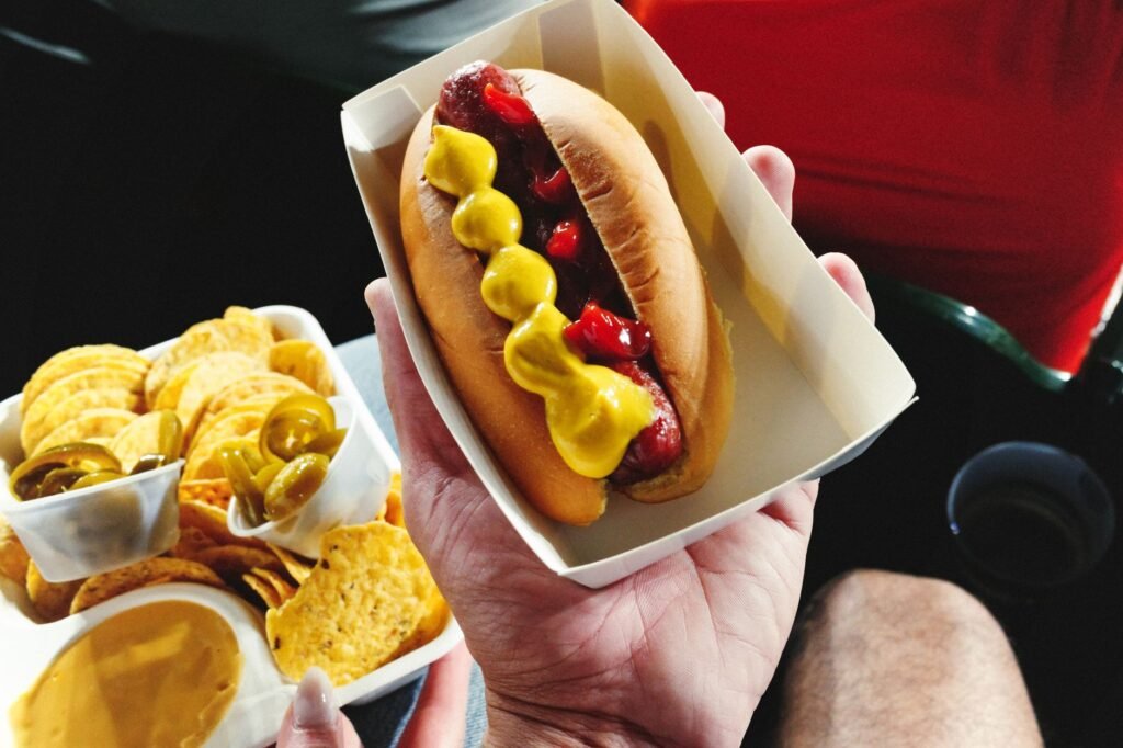 A hand holding a hot dog in a cardboard tray with a thick zigzag of yellow mustard and ketchup along the sausage, beside a tray of tortilla chips with cheese sauce and jalapenos