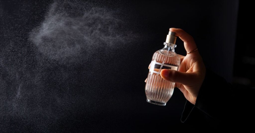 A hand spraying a ribbed glass perfume bottle against a dark background, the fine mist drifting sideways well past where it was aimed