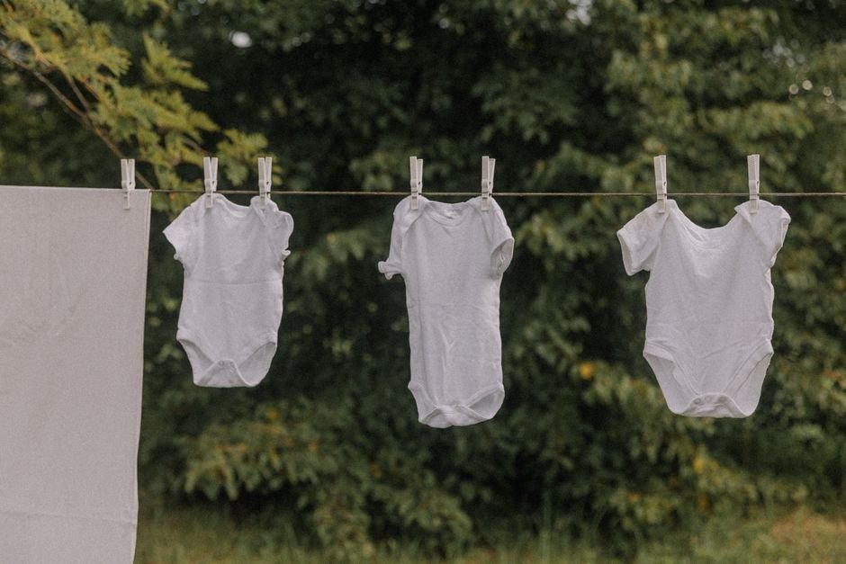 Three white baby bodysuits pegged side by side on an outdoor washing line in front of green trees, with a white sheet hanging alongside