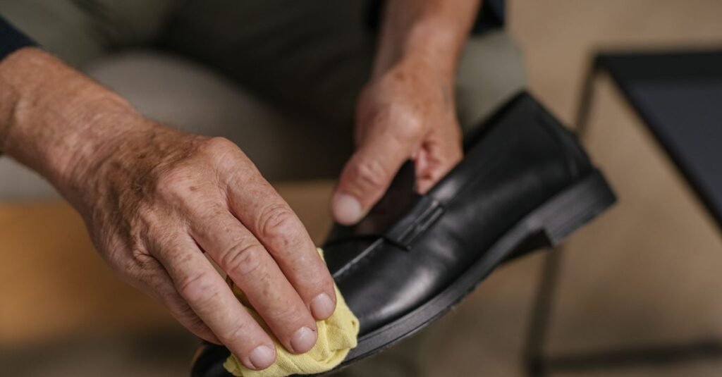 Bare hands buffing a black leather loafer with a yellow cloth, the shoe held inches from the olive trousers the person is wearing