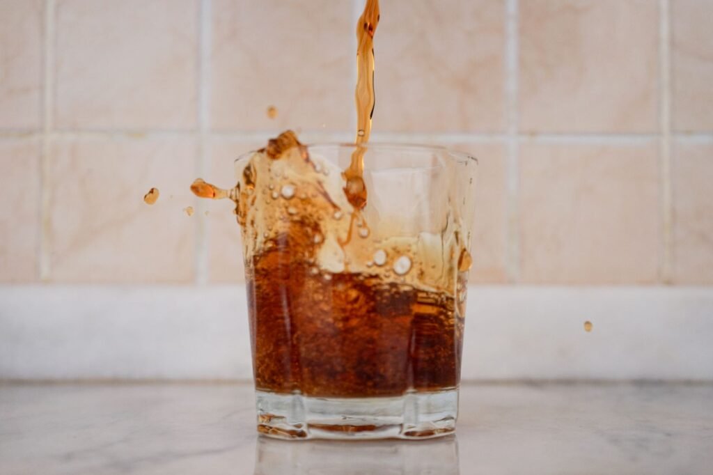 Dark cola being poured into a short tumbler with foam and droplets flying over the rim onto a pale counter