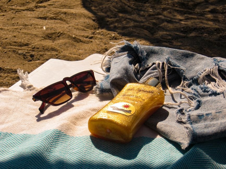 A yellow bottle of sunscreen lying on a striped beach towel next to sunglasses and a pale denim wrap, in strong sunlight on sand