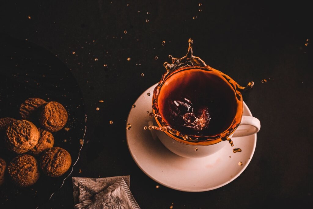 Overhead shot of dark tea splashing up out of a white cup and saucer on a dark table, droplets flying, a used teabag and biscuits alongside