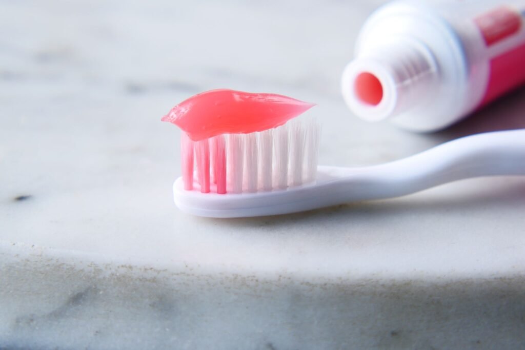 A white toothbrush loaded with a stripe of bright pink toothpaste resting on a marble surface beside an open tube