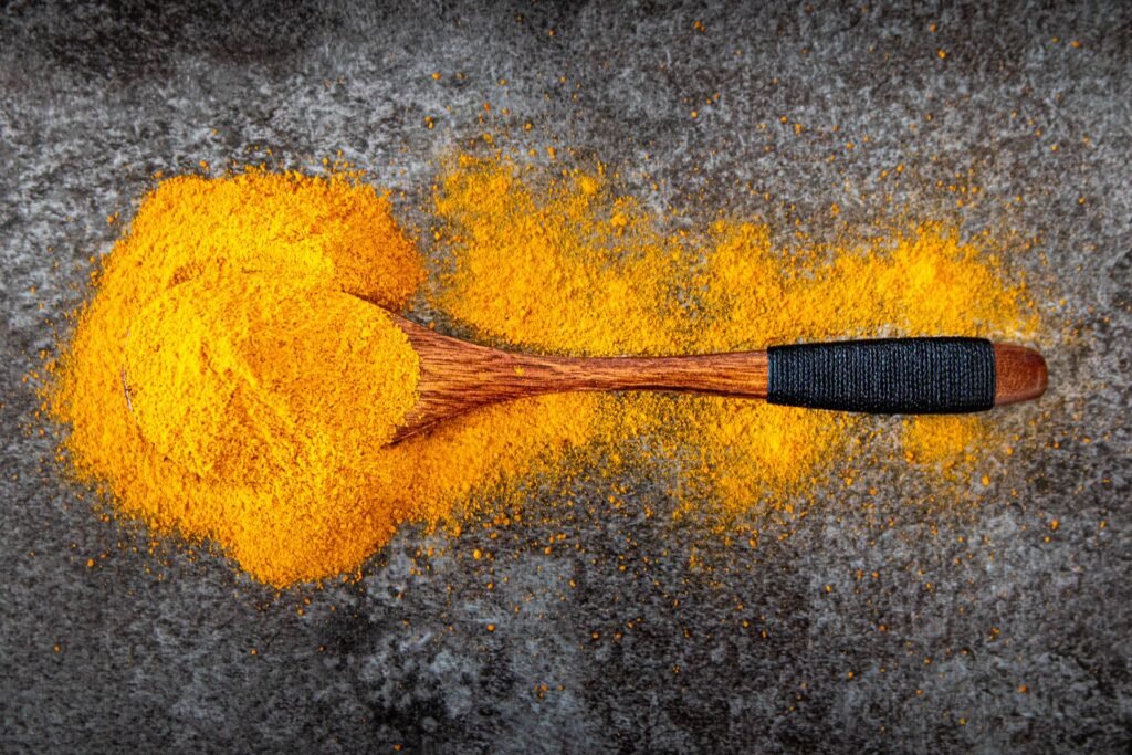 Bright yellow turmeric powder spilled in a heap across a dark grey stone surface with a wooden spoon lying in it