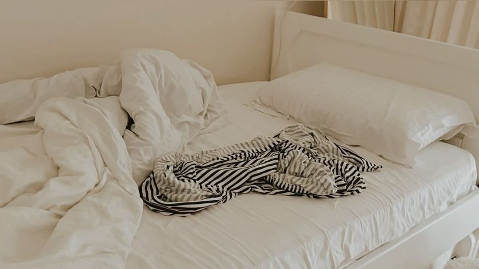An unmade bed with the white duvet pushed back and a black and white striped top dropped on the sheet, waiting to be stripped and washed