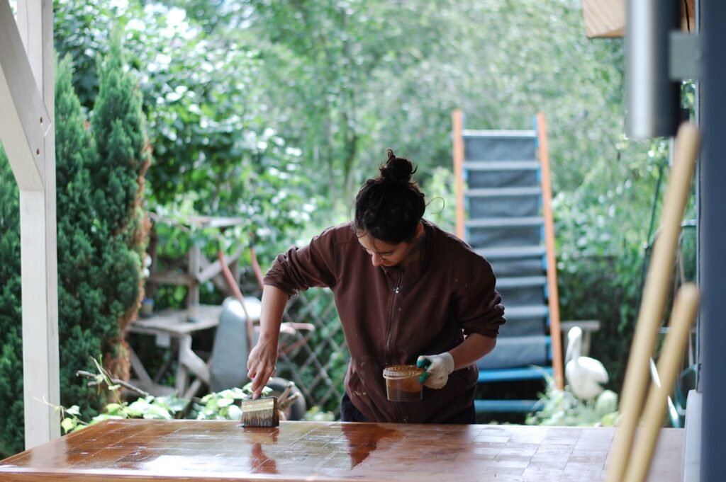 Woman in a dark work jacket brushing wood stain across a large tabletop outdoors while holding the open tin
