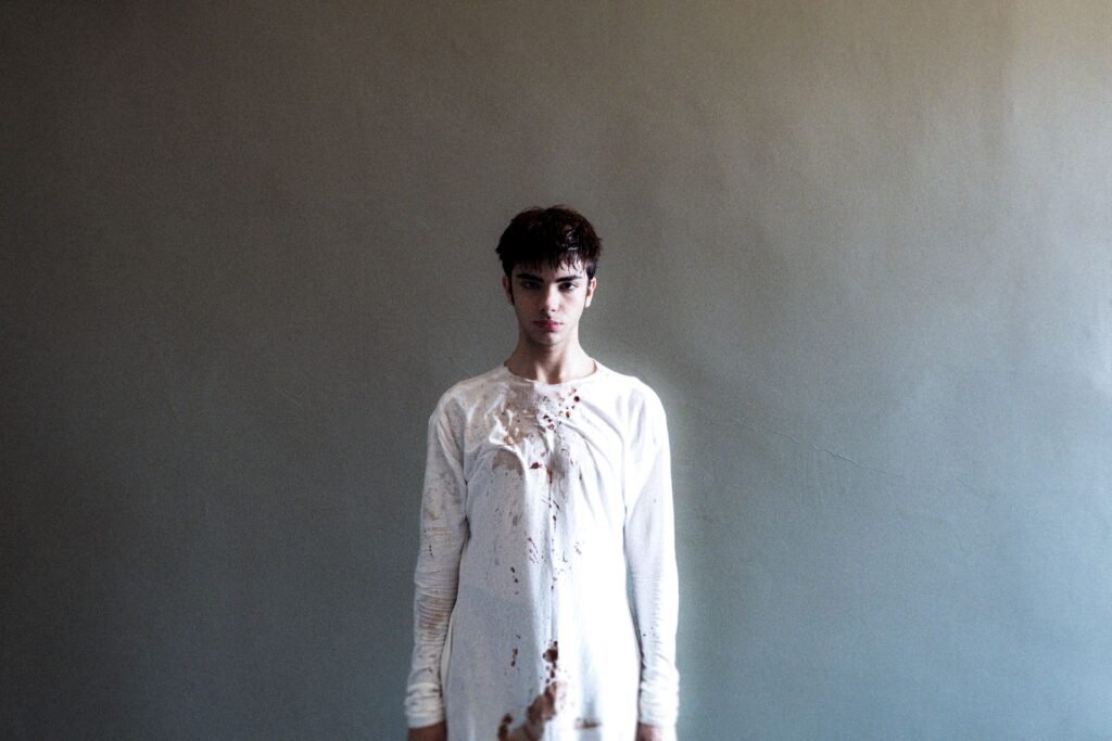 A person standing against a plain grey wall in a white long-sleeved top spattered down the front with dried dark red marks