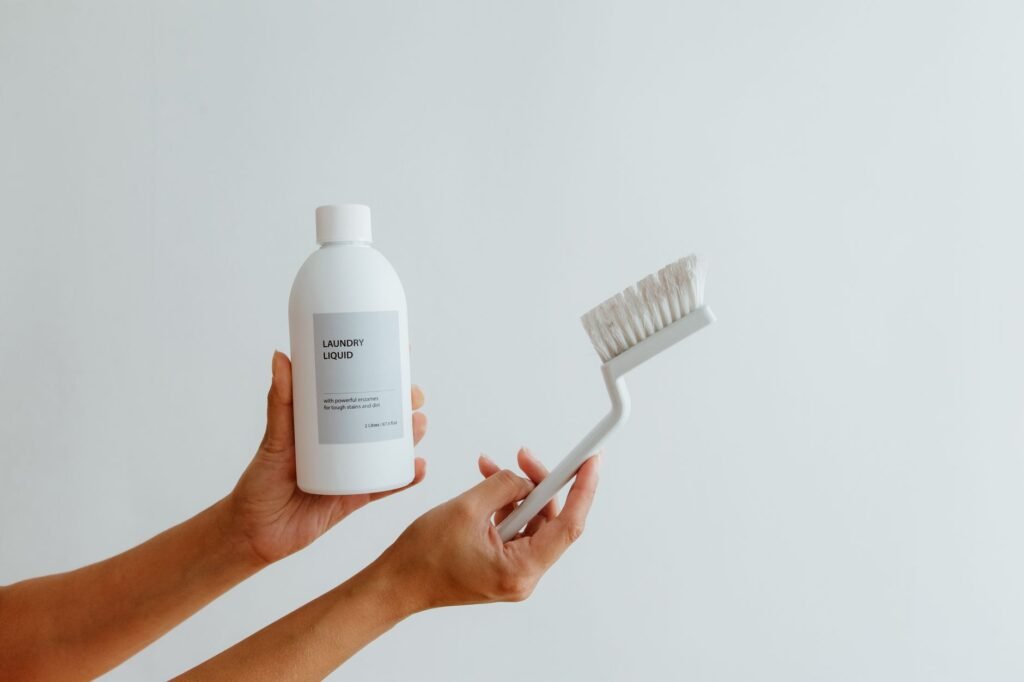 Two hands holding a plain white bottle labelled laundry liquid and a white stiff-bristled scrubbing brush against a pale grey wall