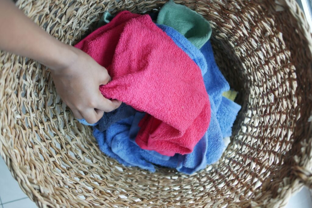 A hand lifting a bright pink towel out of a woven basket packed with blue, green and pink coloured laundry