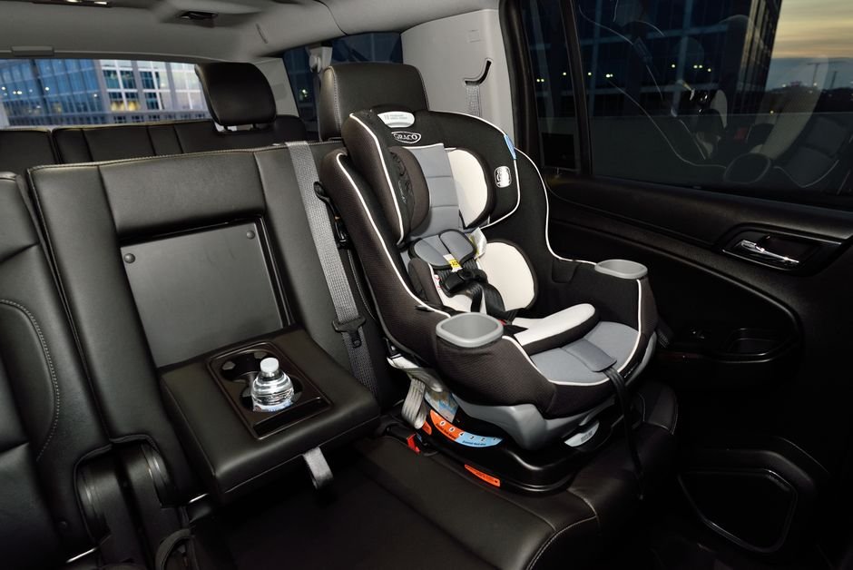 A grey and black convertible child car seat strapped into the rear leather seat of a car, its five-point harness and chest clip lying across the padded cover