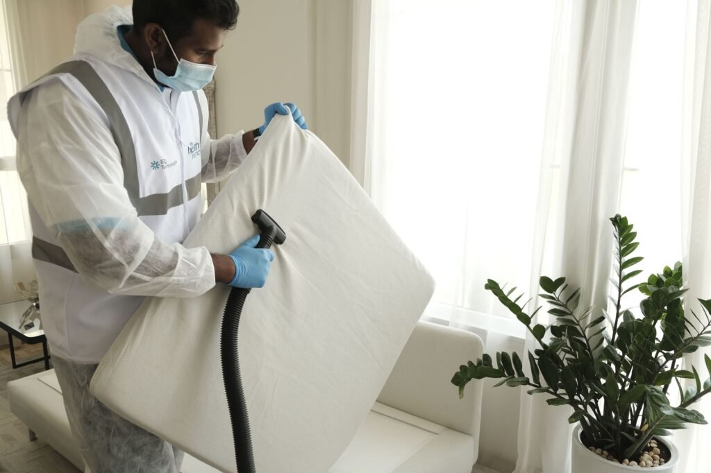 A technician in coveralls, mask and gloves holding up a thin white foam mattress pad and running a vacuum extraction nozzle across it