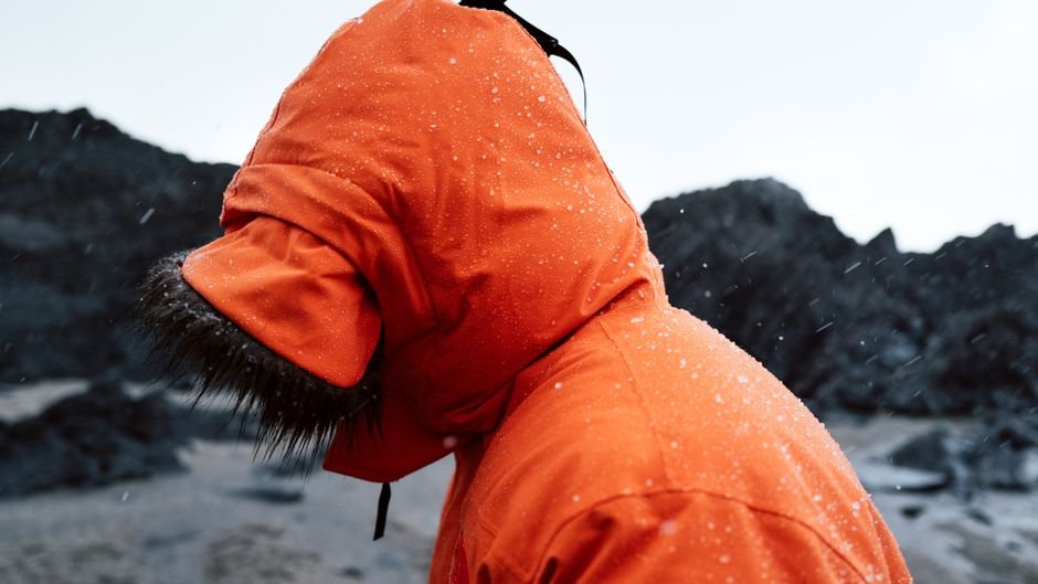 Side view of a hooded orange waterproof jacket in sleet, with water standing in beads across the shoulder and hood rather than soaking in