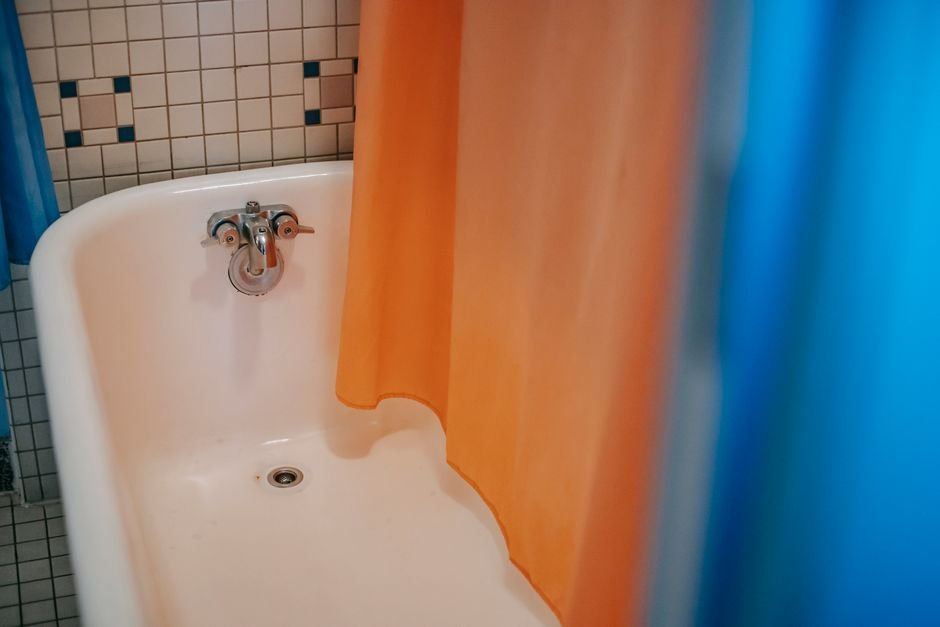 An orange shower curtain hanging down inside a white enamel bathtub with a blue outer curtain beside it, the hem sitting where water collects