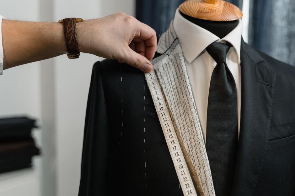 Tailor holding a tape measure against a part-made jacket front, with white basting stitches and the pale interlining strip visible under the lapel