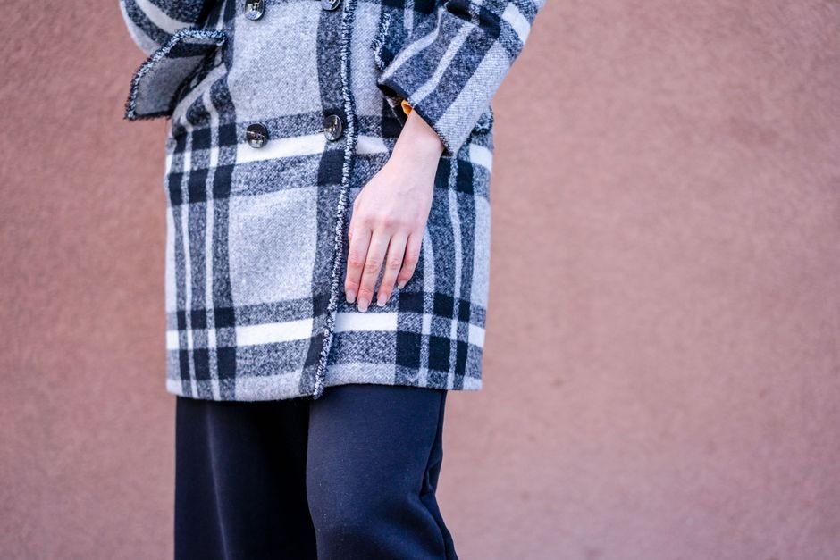 Close-up of a heavy grey and black checked wool coat with raw fringed seam edges and dark horn buttons, worn against a pink wall