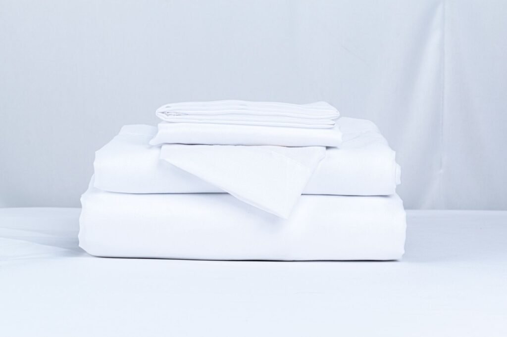 A stack of folded white bed linens — flat sheet, fitted sheet and pillowcases — on a pale surface
