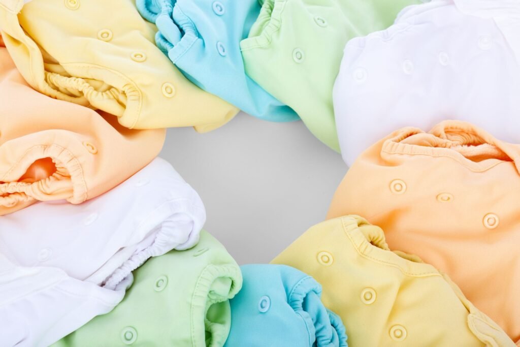 Overhead view of pastel yellow, blue, green, peach and white cloth diaper covers with press-stud snaps and elasticated legs, arranged in a ring on a grey background