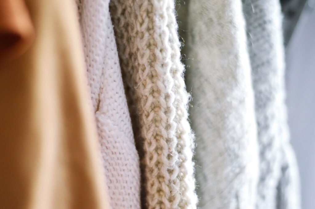 Close-up of four knitted sweaters hanging side by side in cream, white and pale blue, the chunky cable stitch and loose fibre ends lit by soft daylight