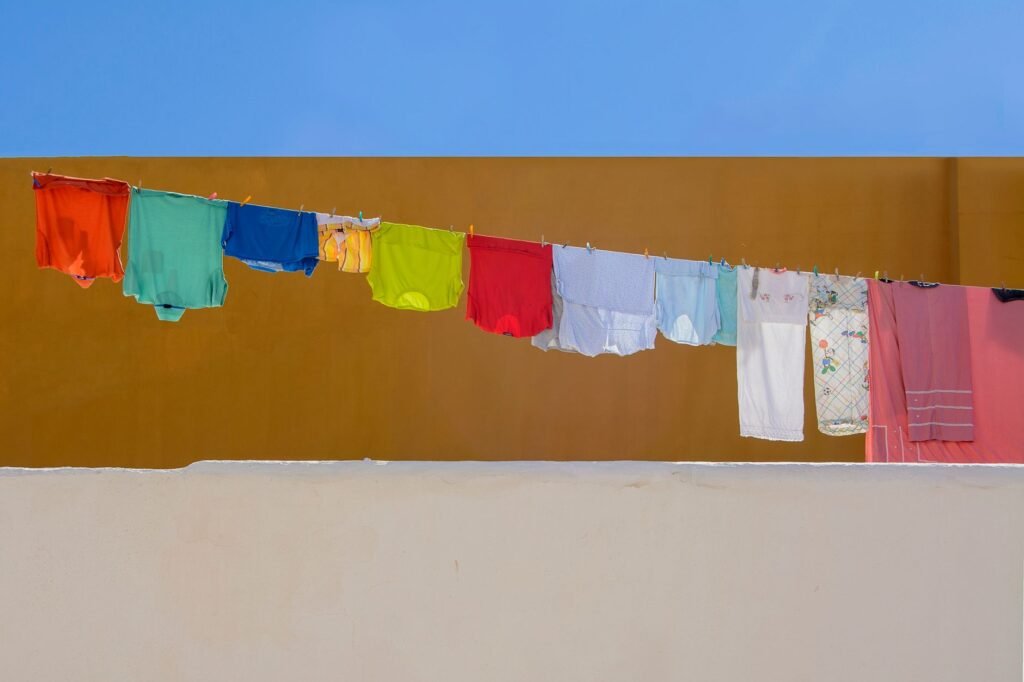 A washing line strung across an ochre wall under a blue sky, hung with a red, teal, blue, yellow and green row of garments running into pale blues, whites and a pink sheet at the end